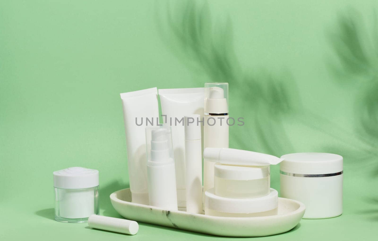 A collection of various white cosmetic containers, including tubes, jars, and bottles, are arranged on a white stand against a light green background with leaf shadows.
