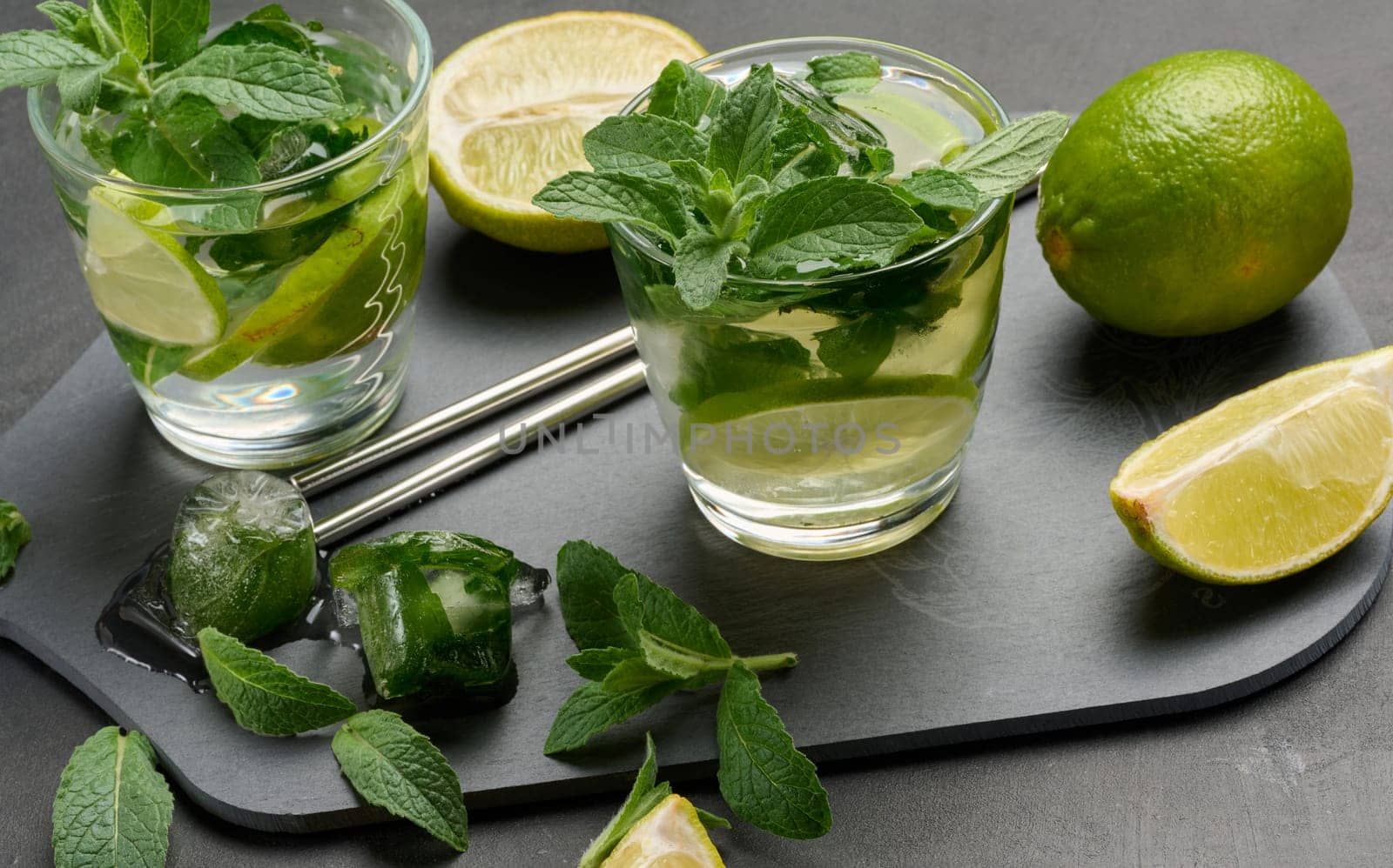 Glass of mojito cocktail with fresh mint leaves, lime slices, and ice cubes on a dark stone background. Refreshing summer drink with stainless steel straws
