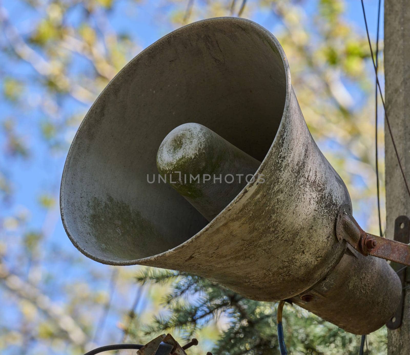 An old, dirty, grey loudspeaker is mounted on a pole. In the background, there is a blurred image of the sky and tree branches. View from below.