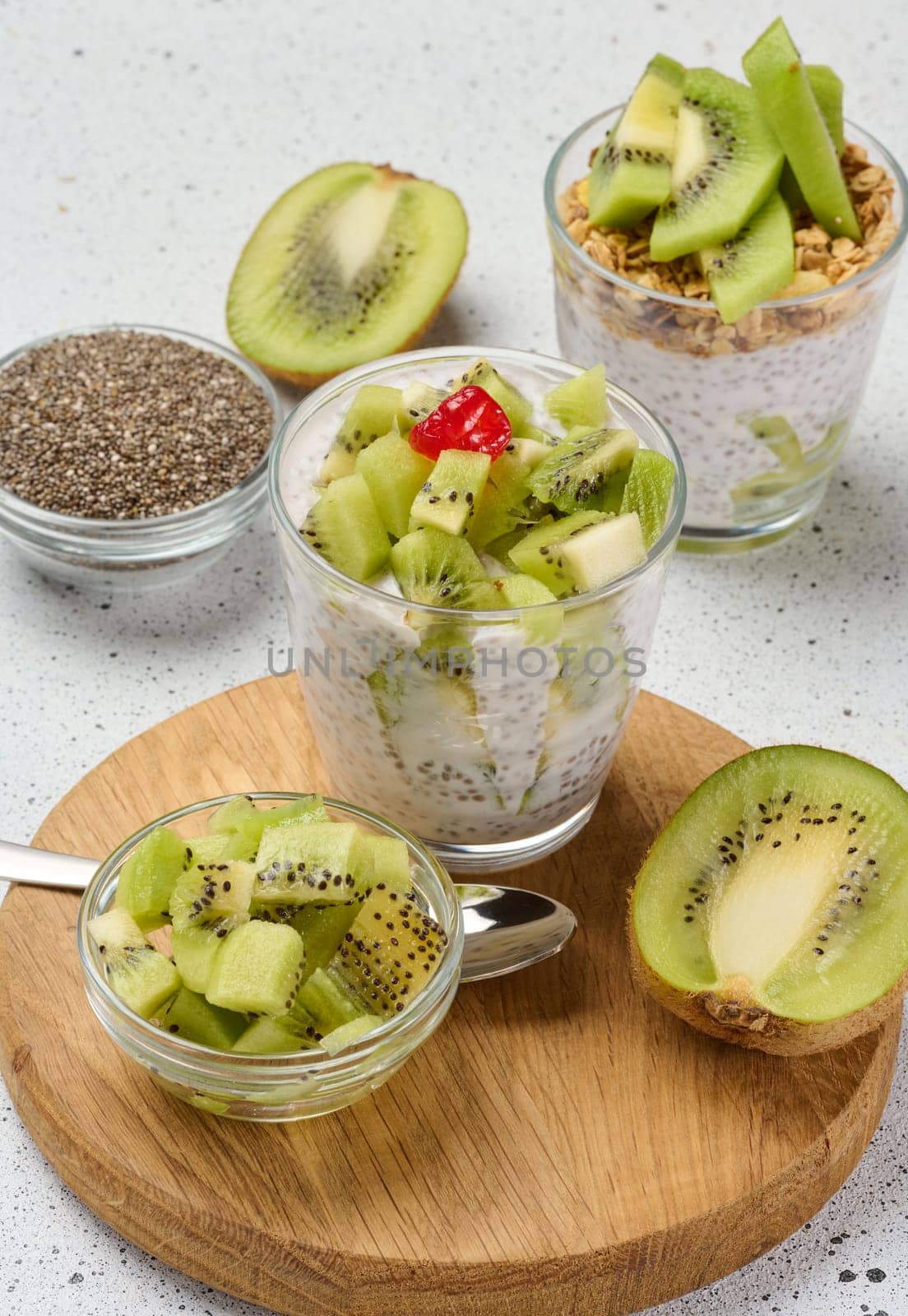 A glass of chia seed pudding layered with kiwi and granola, topped with kiwi slices, sits on a wooden board with ingredients around it
