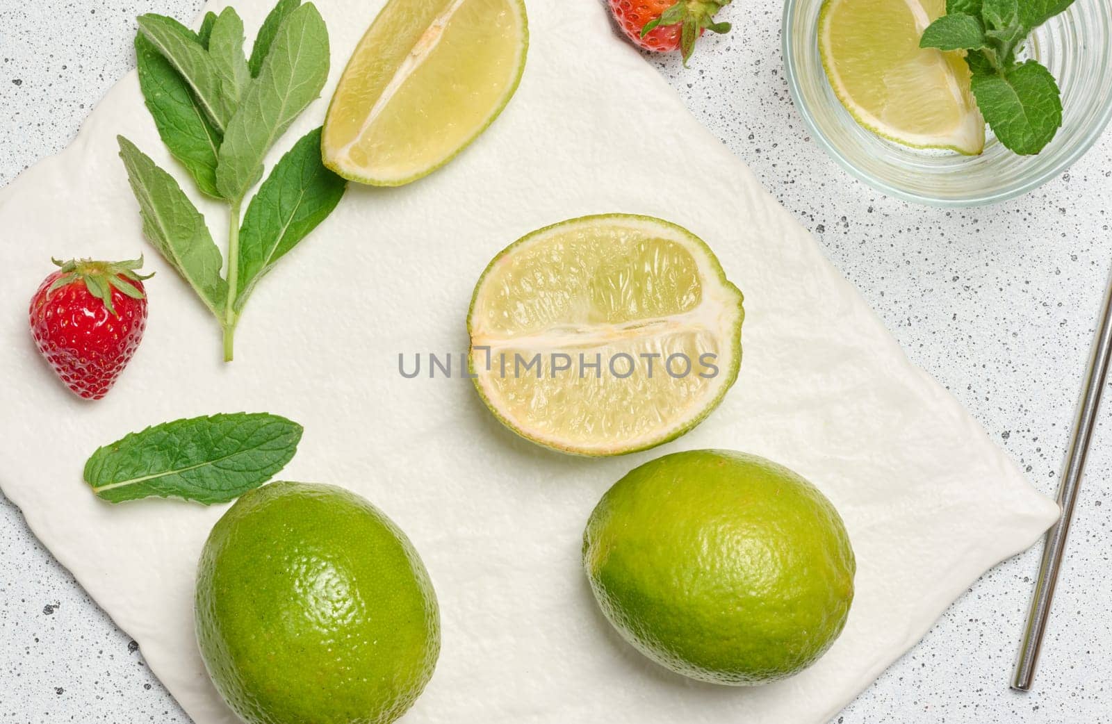 A top down view of ingredients for a refreshing drink, including whole and sliced limes, fresh mint sprigs, empty glass, metal straw