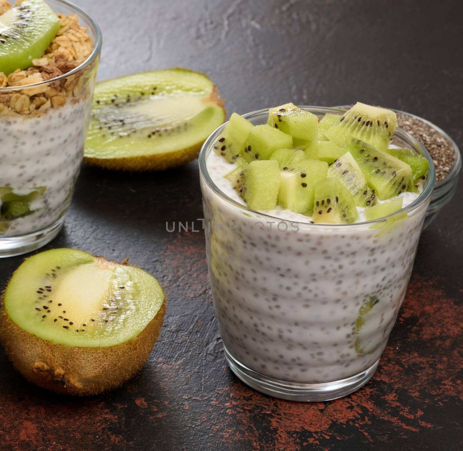 Two glasses of chia seed pudding, one topped with diced kiwi and the other with granola and banana chips, are on a dark surface with ingredients