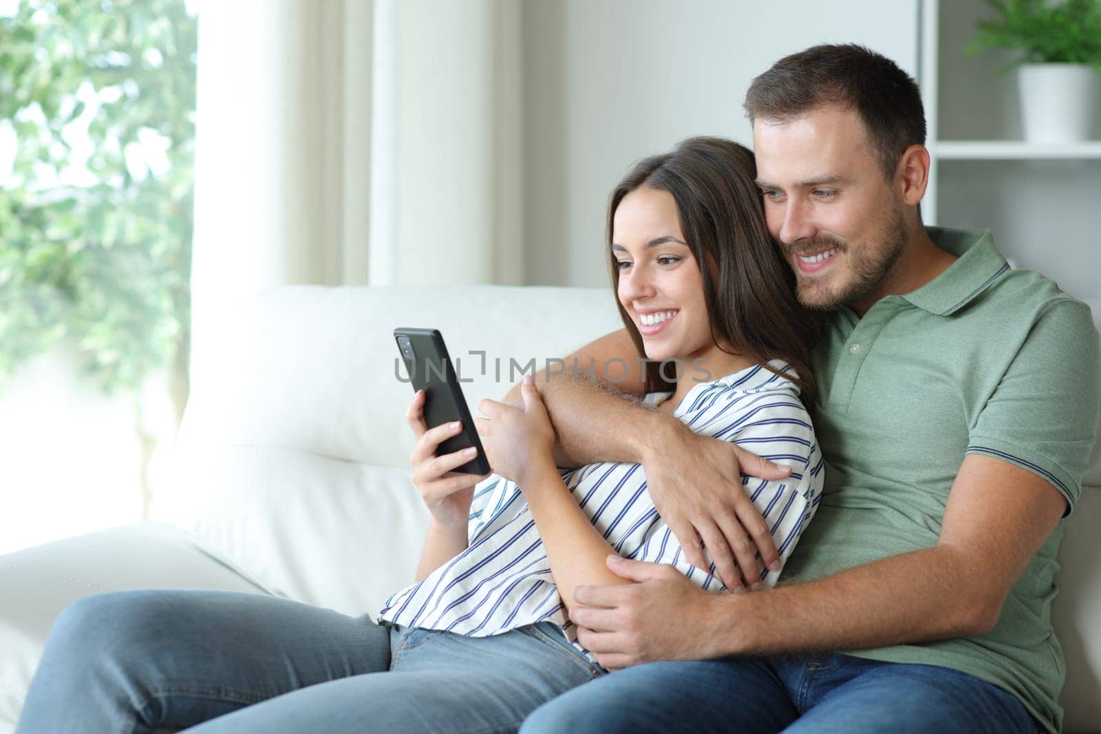 Happy couple using mobile phone together sitting ona couch at home