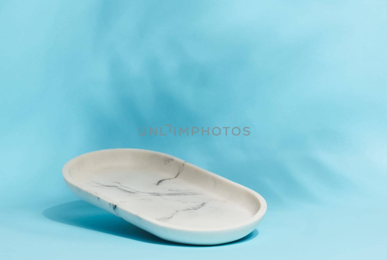 An empty, oval shaped soap dish made of white marble with gray veining sits on a light blue background with soft, blurred leaf shadows