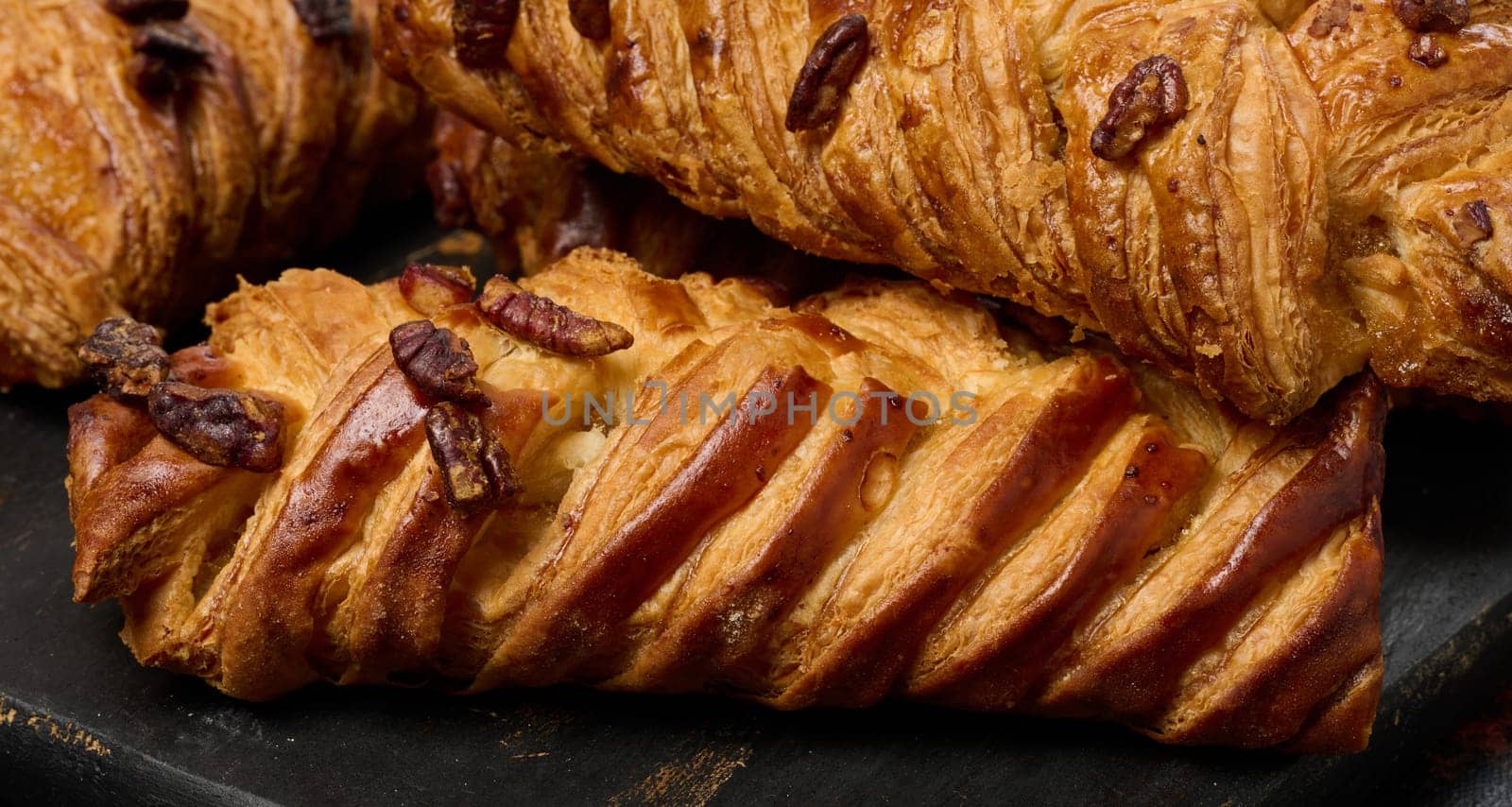 Golden brown maple pecan braid pastries are arranged on a dark, textured surface. Braided dough is flaky and topped with pecans