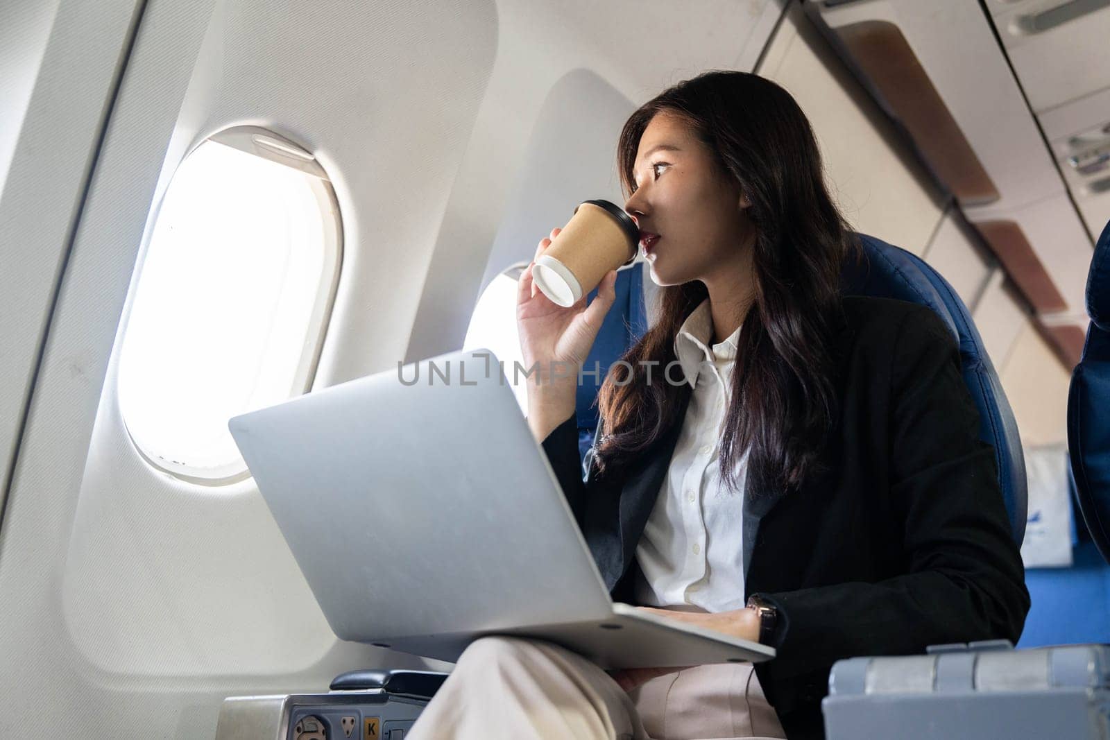 A businesswoman enjoying a coffee while using her laptop on an airplane, illustrating a modern approach to work during travel.