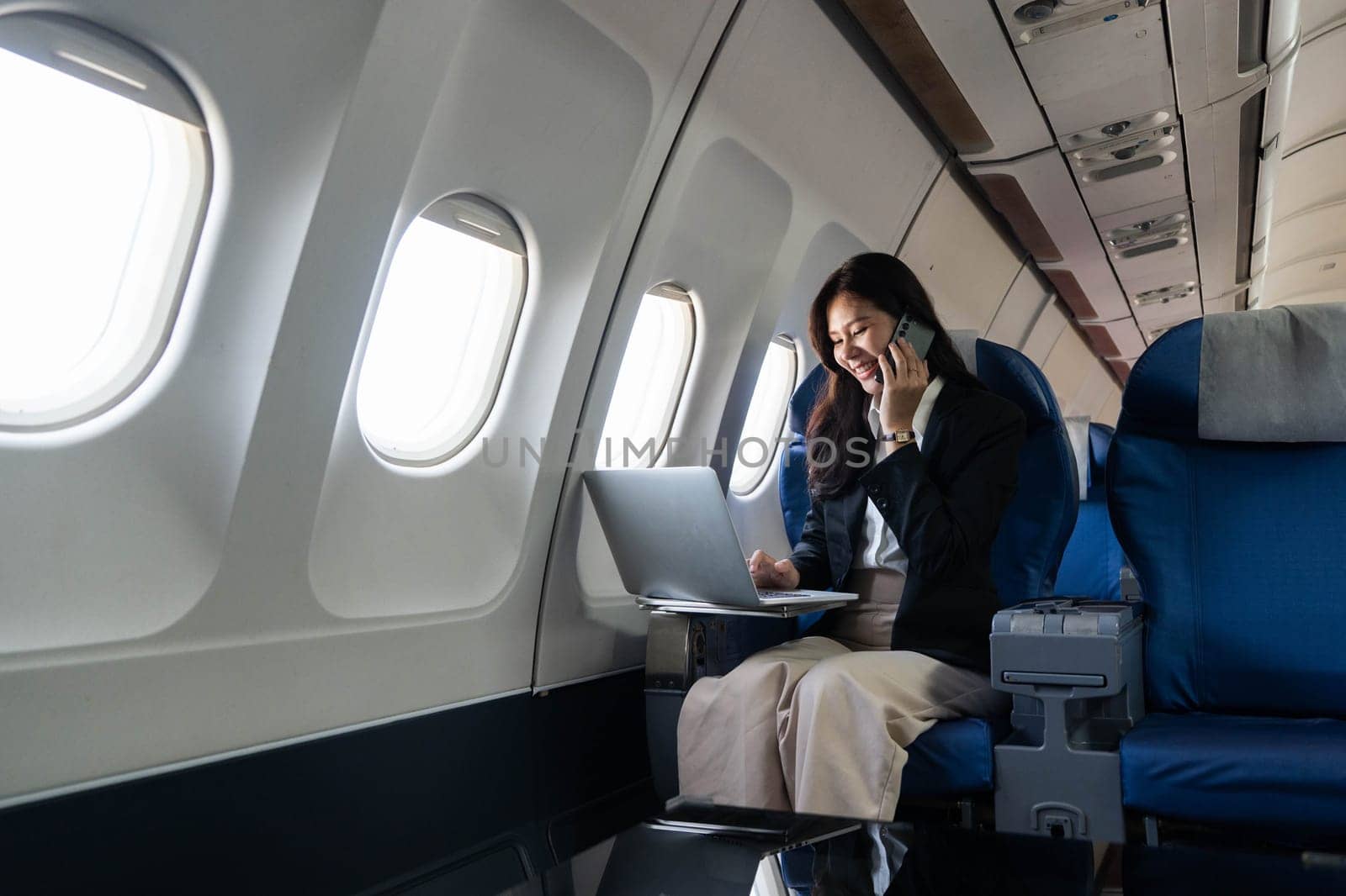 A businesswoman multitasking on a flight, engaged in phone conversation while working on her laptop, embodying the essence of modern travel.