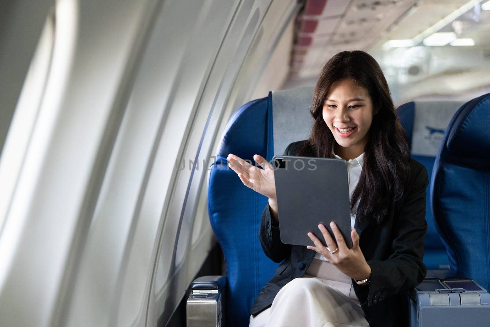 A young businesswoman joyfully waves at her tablet camera while seated on an airplane, connecting during her trip.