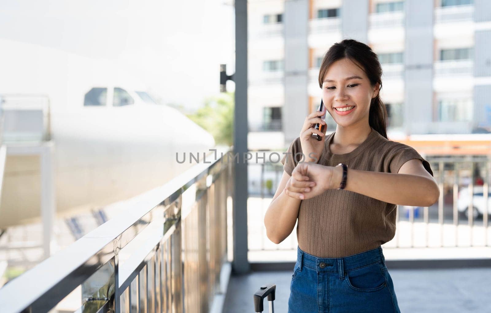 A young woman smiles while talking on her phone, standing by her luggage at the airport, feeling excited for her upcoming journey.