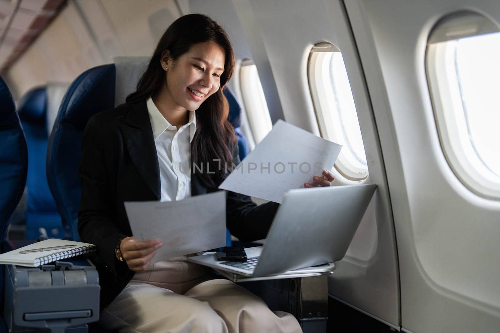 A happy professional woman smiling while reviewing documents and working on her laptop during a flight, representing positivity in business travel.