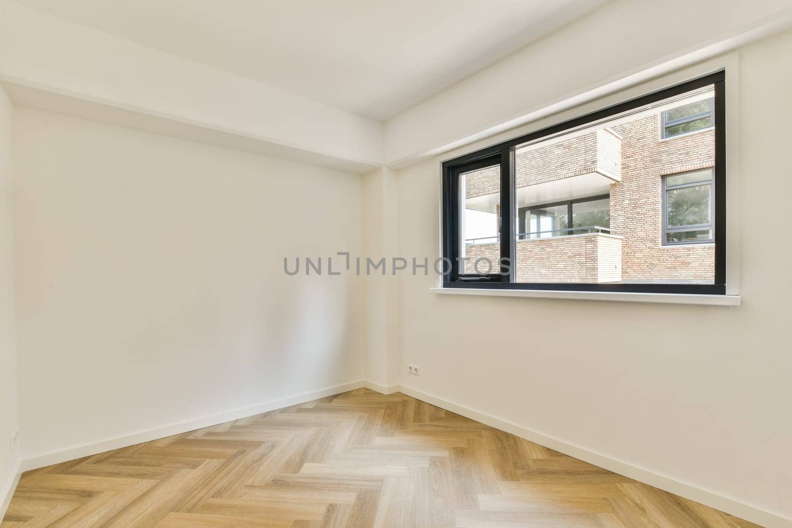 A spacious modern room featuring large windows that allow natural light to flood in. The wooden flooring adds warmth to the minimalist design.
