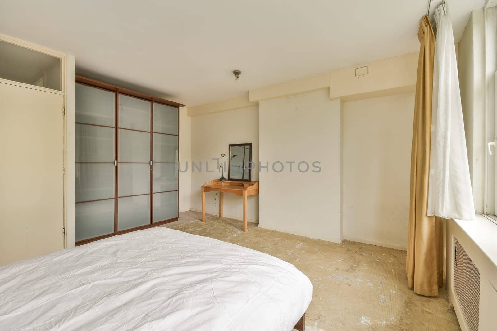 A spacious minimalist bedroom featuring a bed, a wooden table, a mirror, and large windows allowing natural light to flood the room. Neutral tones enhance serenity.
