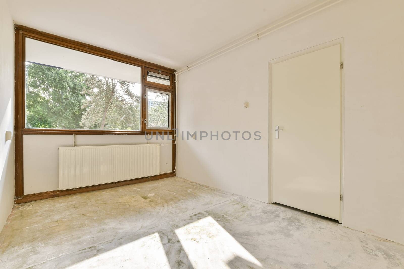 A bright, empty room featuring a large window that allows natural light to flood in, enhancing its spacious feel. The walls are plain, and the floor is unfinished.
