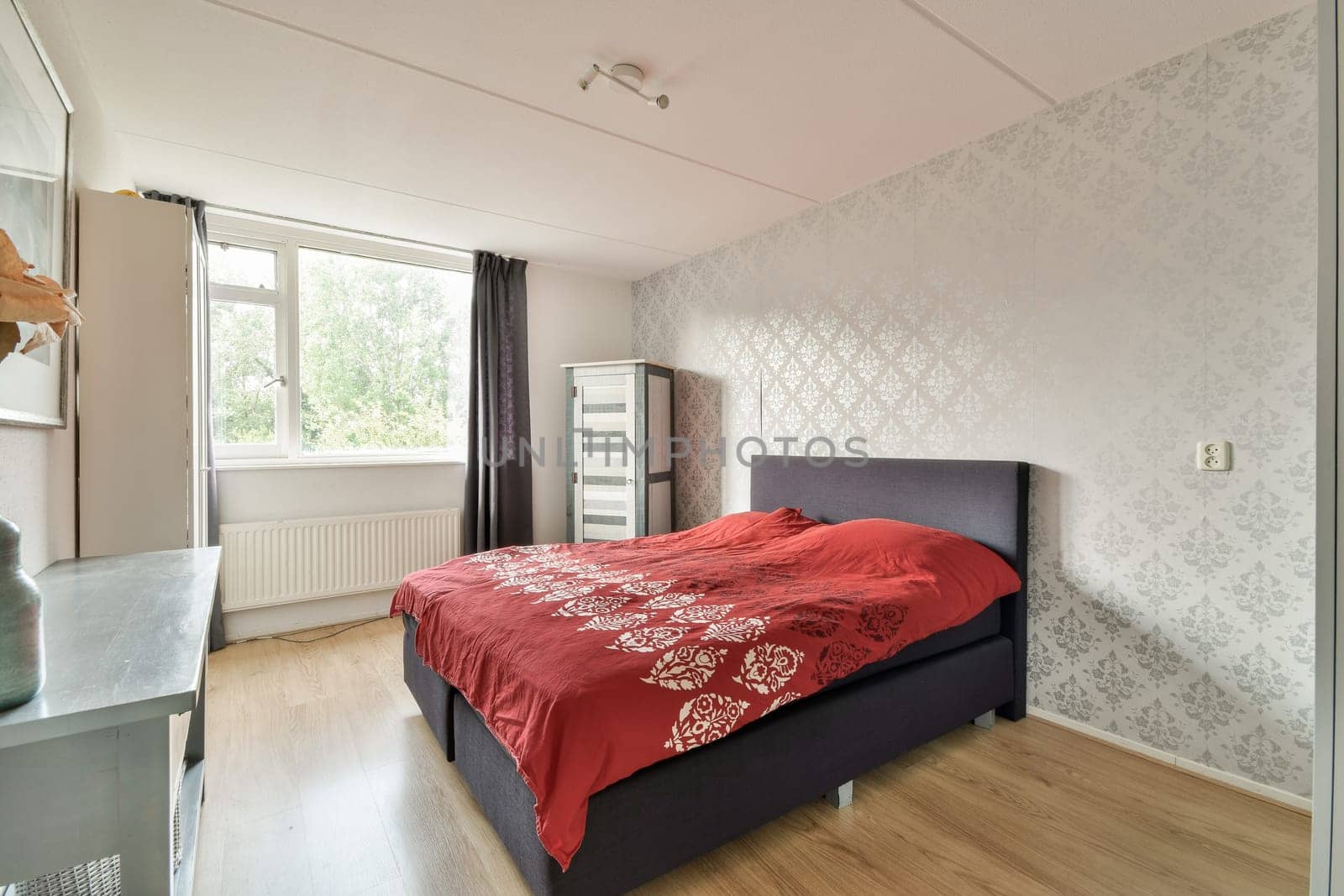 A stylish modern bedroom featuring a cozy red bedspread, wooden flooring, and large windows that bring in natural light.