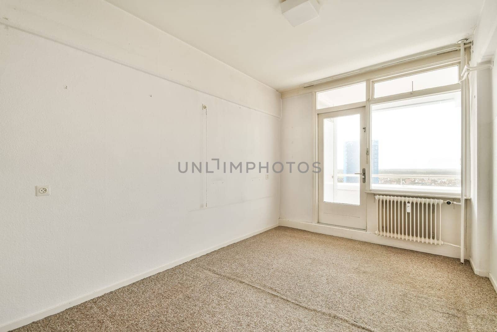 A spacious, minimalistic empty room featuring large windows that allow natural light to flood in, creating a serene atmosphere. The walls are white, enhancing the airy feel.