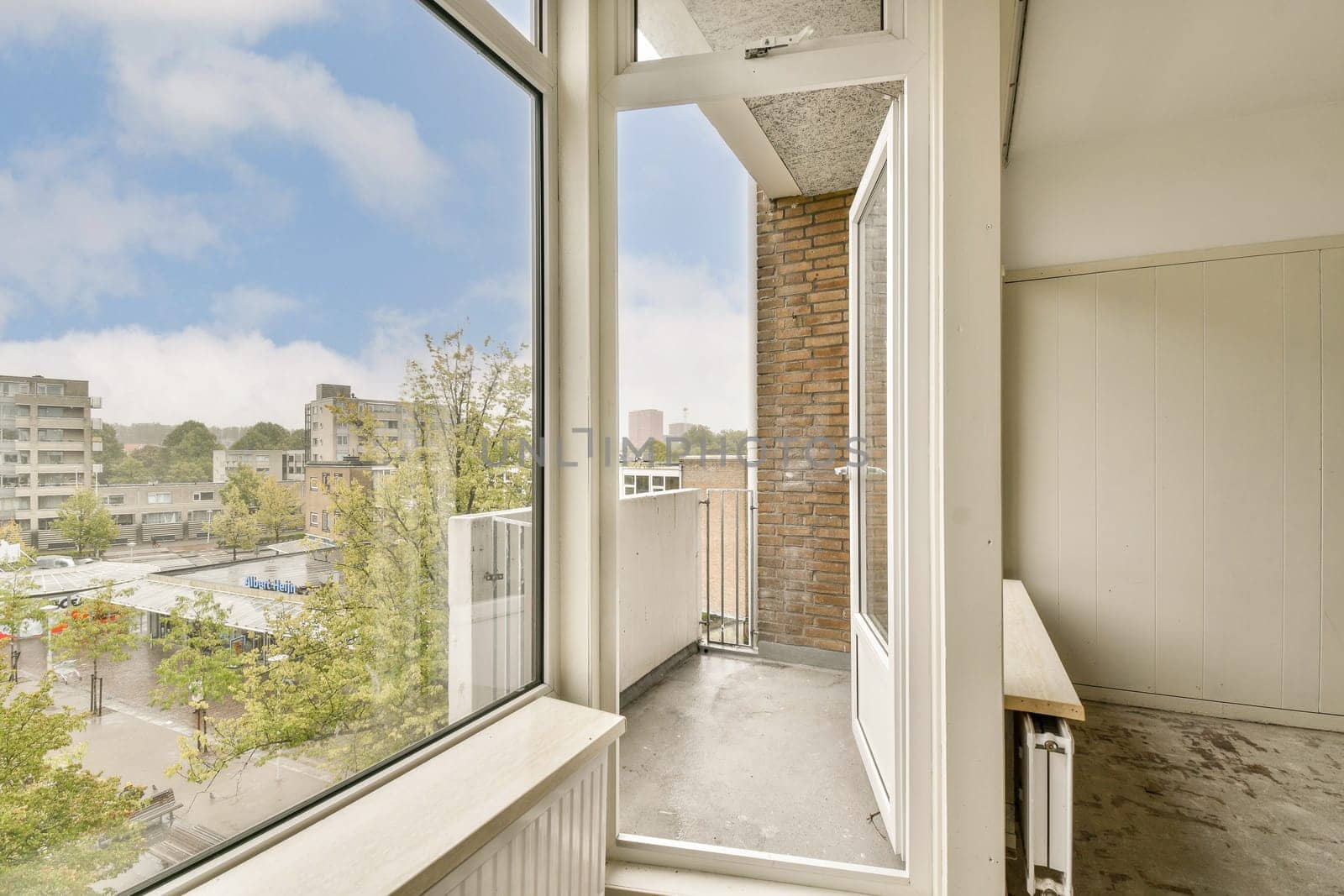 A modern interior view showcasing large windows, allowing natural light to flood a simple balcony space with urban greenery in sight.