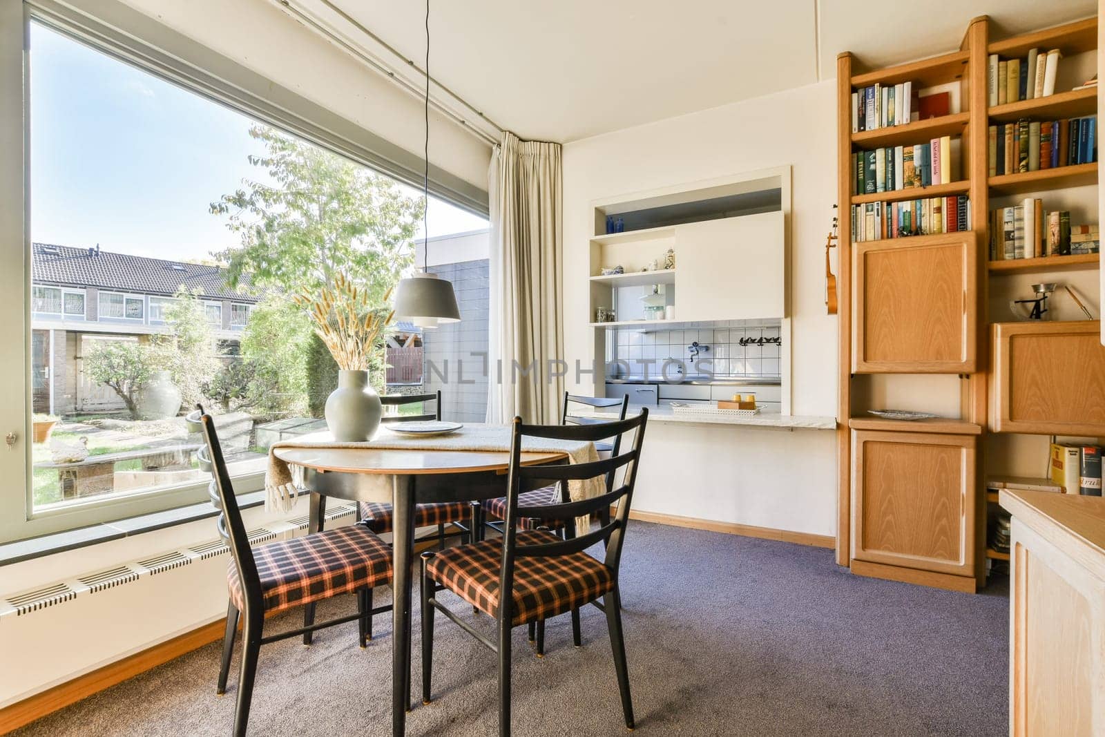 A well-lit dining space featuring a round table with checkered upholstery, surrounded by bookshelves and a view of greenery outside.
