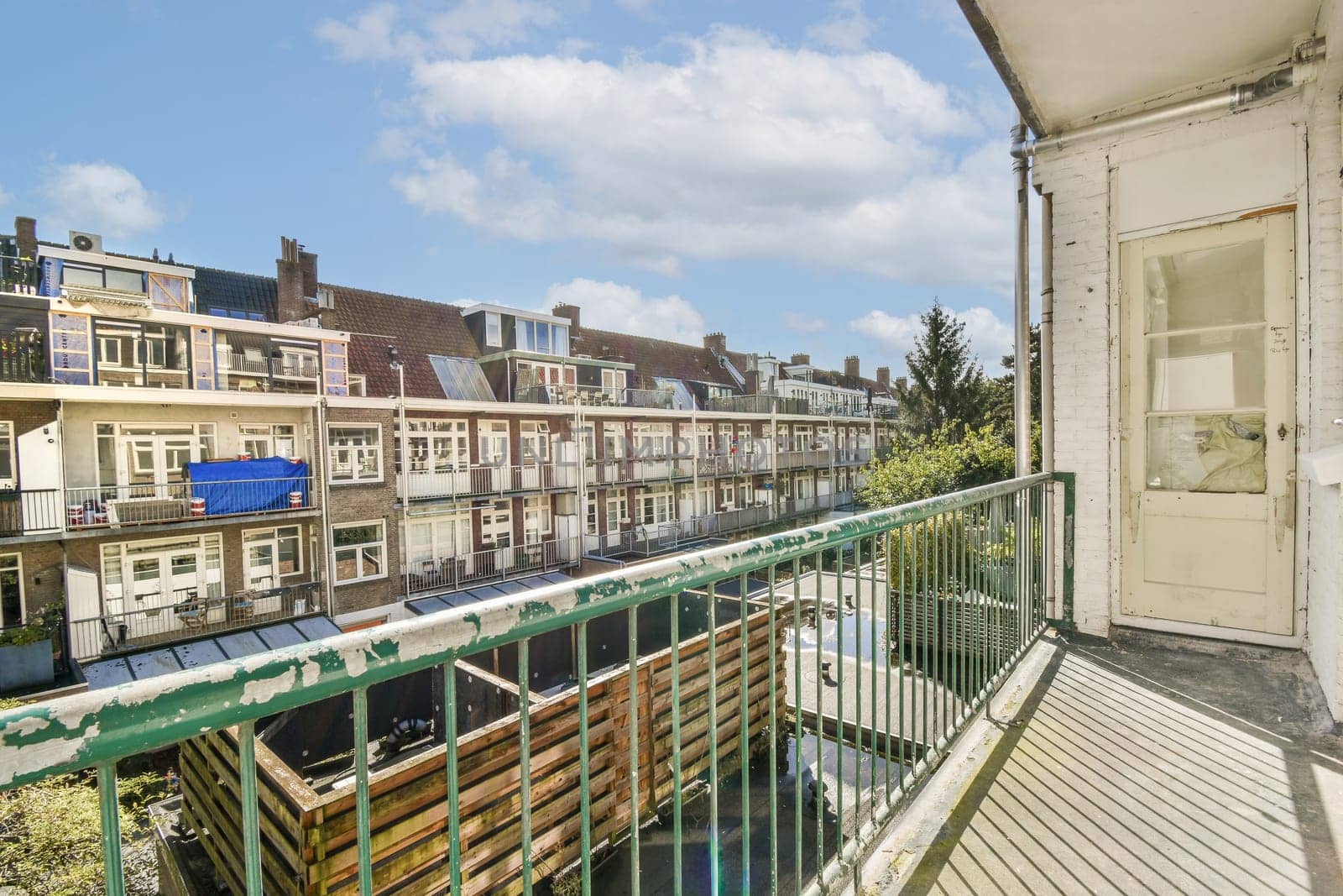 A scenic view showcasing a balcony with railings that overlooks a row of urban buildings under a blue sky with clouds. The greenery adds a touch of nature.