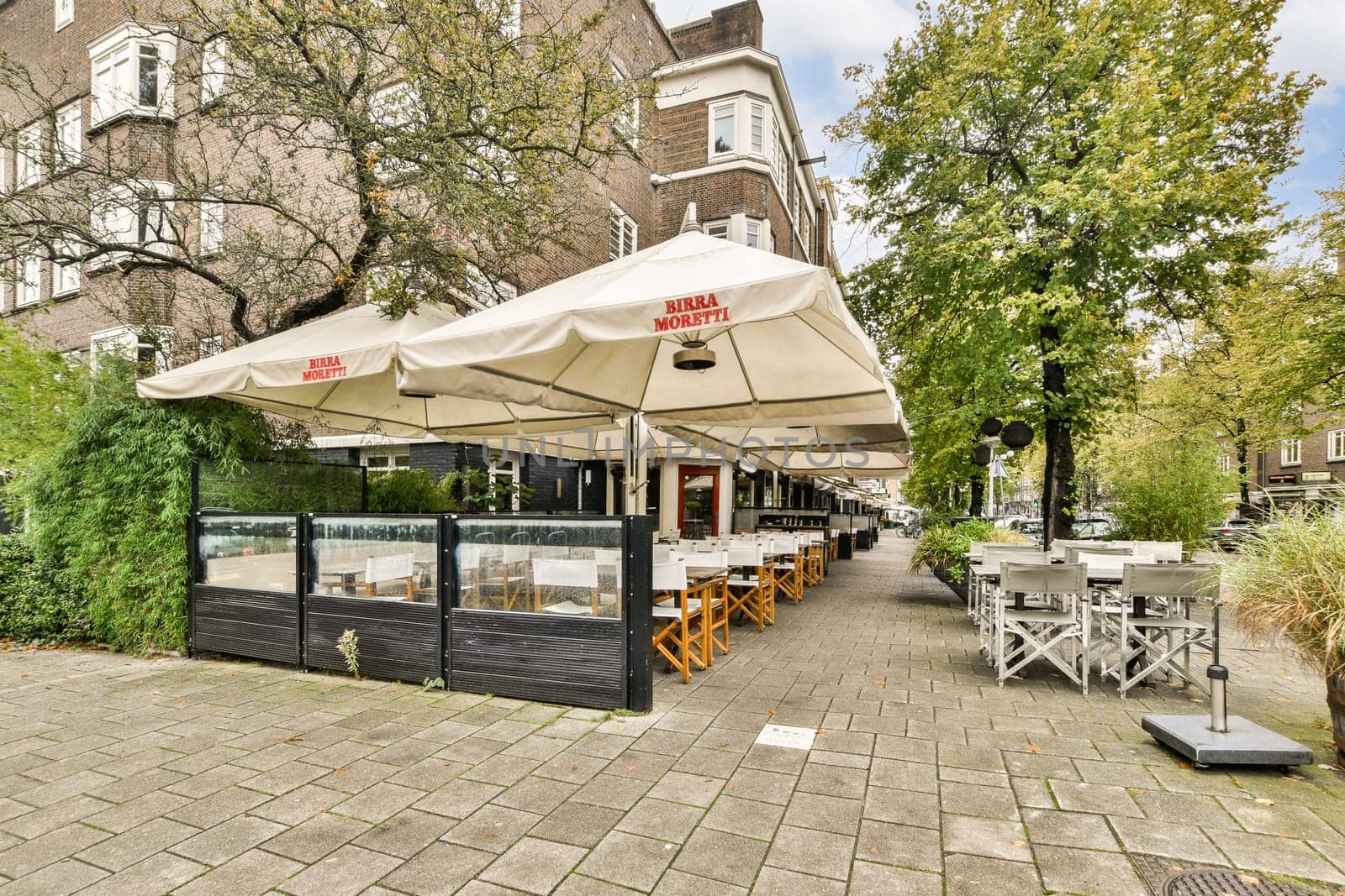 A beautifully arranged outdoor seating area with stylish furniture and shaded umbrellas, surrounded by greenery in an urban setting.