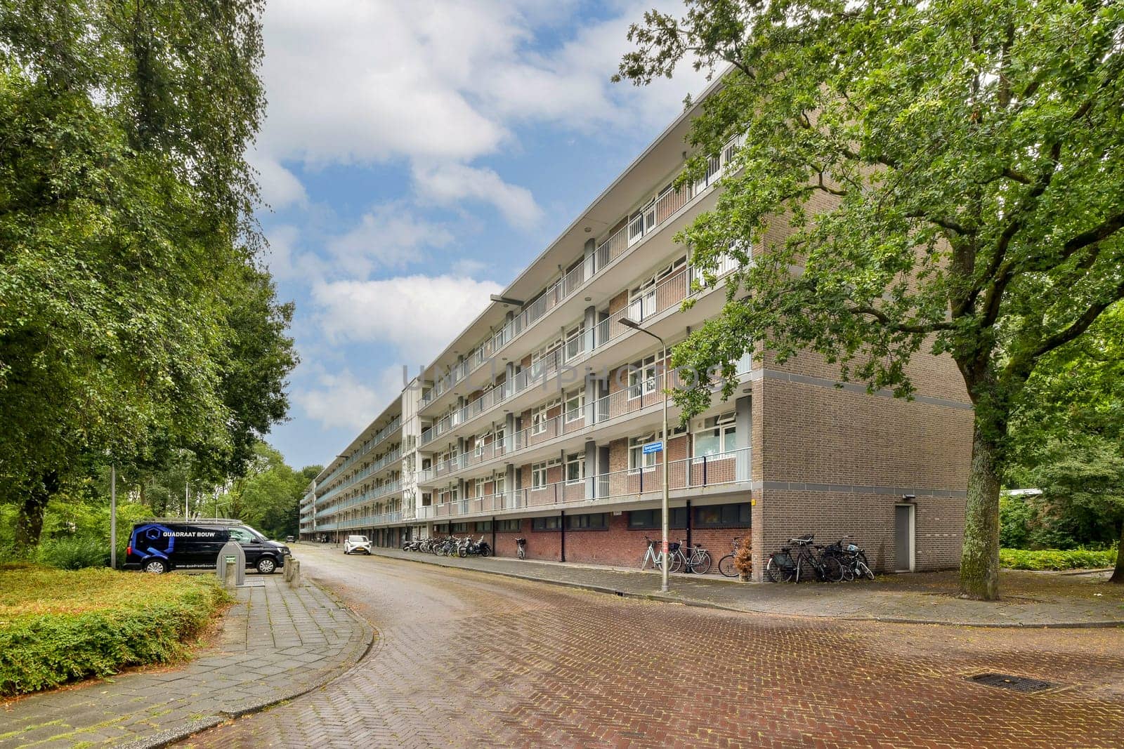 A contemporary residential block viewed from a street, surrounded by trees and parked bicycles, under a cloudy sky that adds depth to the scene.
