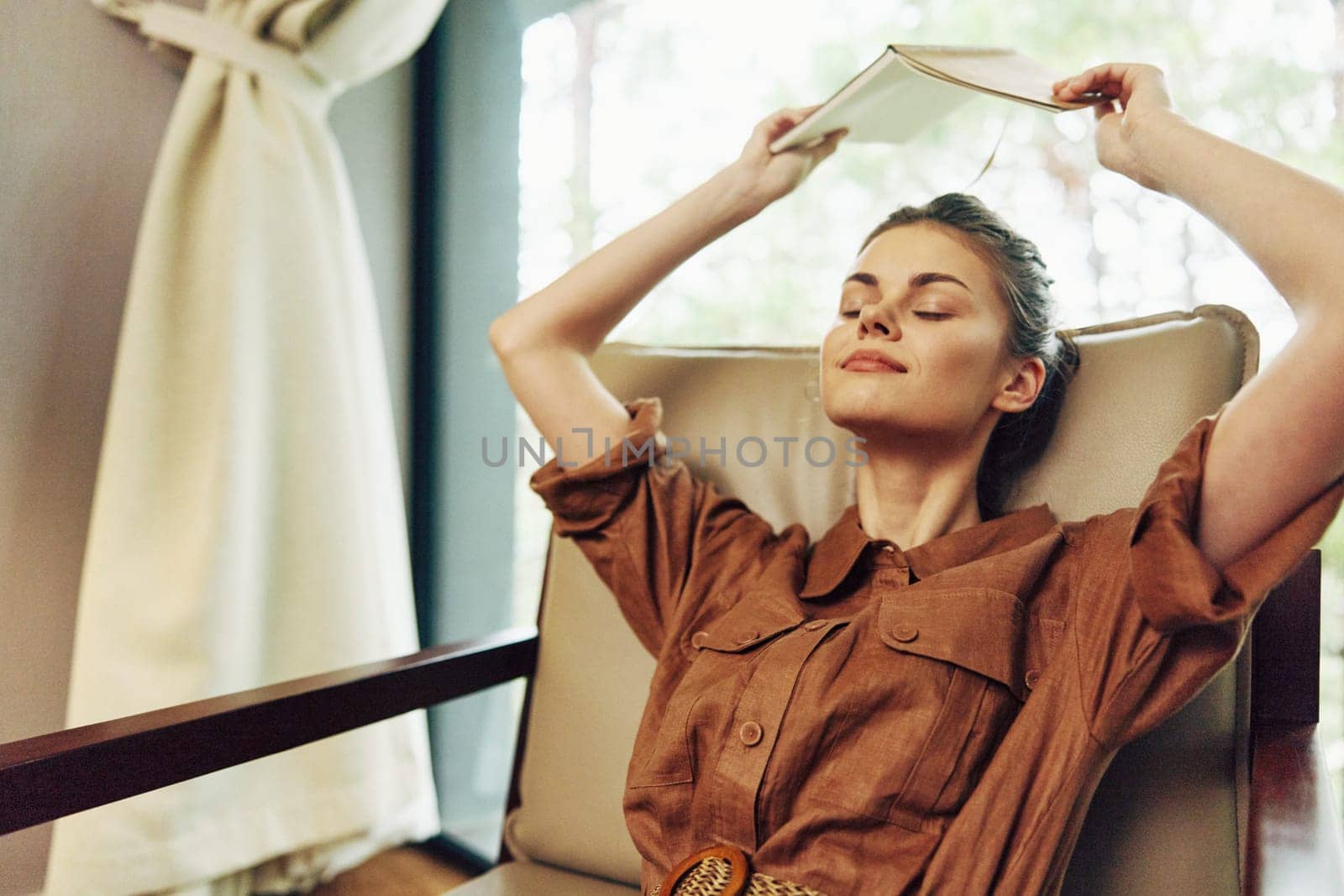 A serene young woman, dressed in a stylish brown shirt, is blissfully relaxing with a book in a cozy, sunlit living room, capturing a sense of calm and comfort