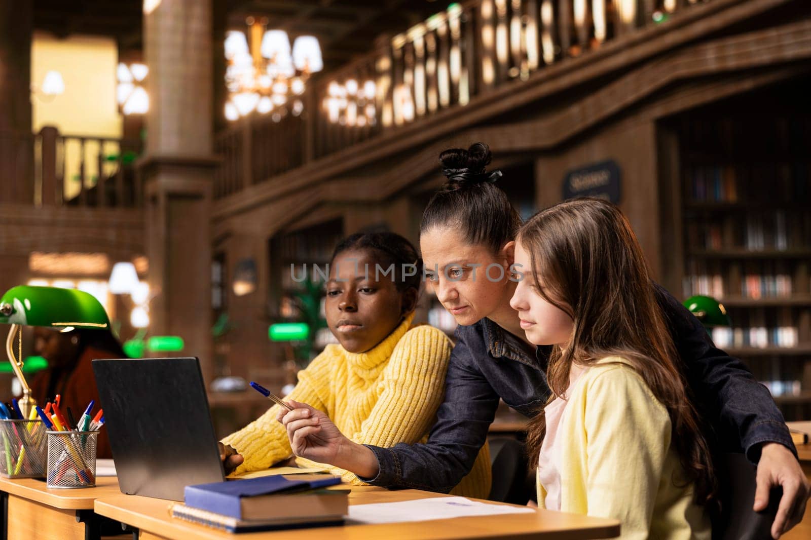 Caucasian tutor guiding two elementary school girls at a library desk, providing private lessons to aid in their academic development with work books and notes. Teacher discussing with scholars.