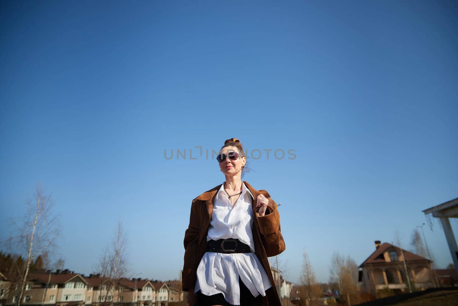 A fashionable woman strolls with confidence in a sunny urban setting, showcasing her unique style against the vivid blue sky.