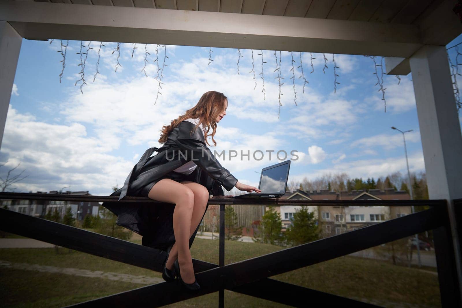 A woman in a black dress sits on a balcony railing using her laptop as clouds drift overhead in a suburban setting.