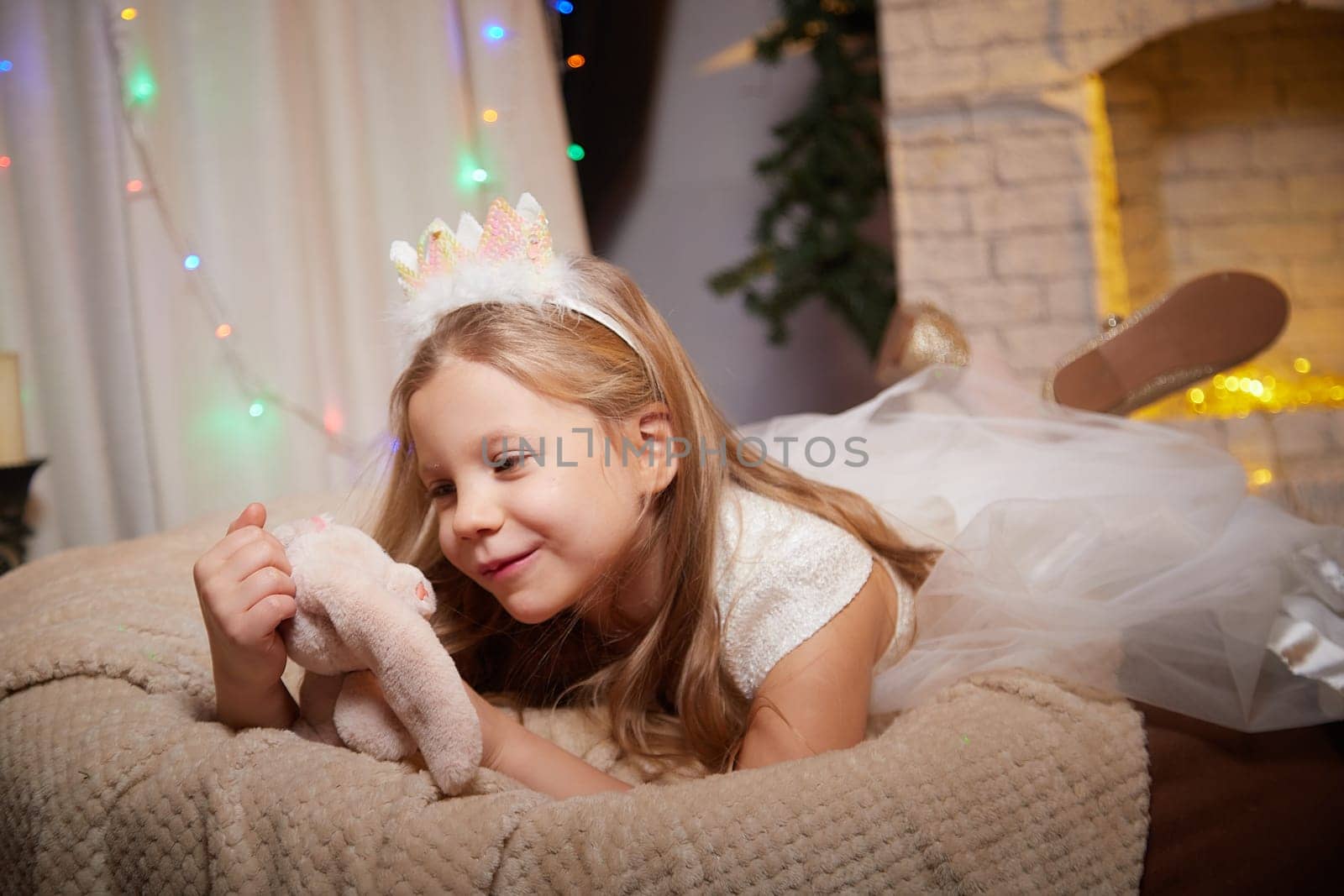 A cheerful child in a white dress and crown plays with a stuffed animal, surrounded by warm holiday decorations in a cozy room.