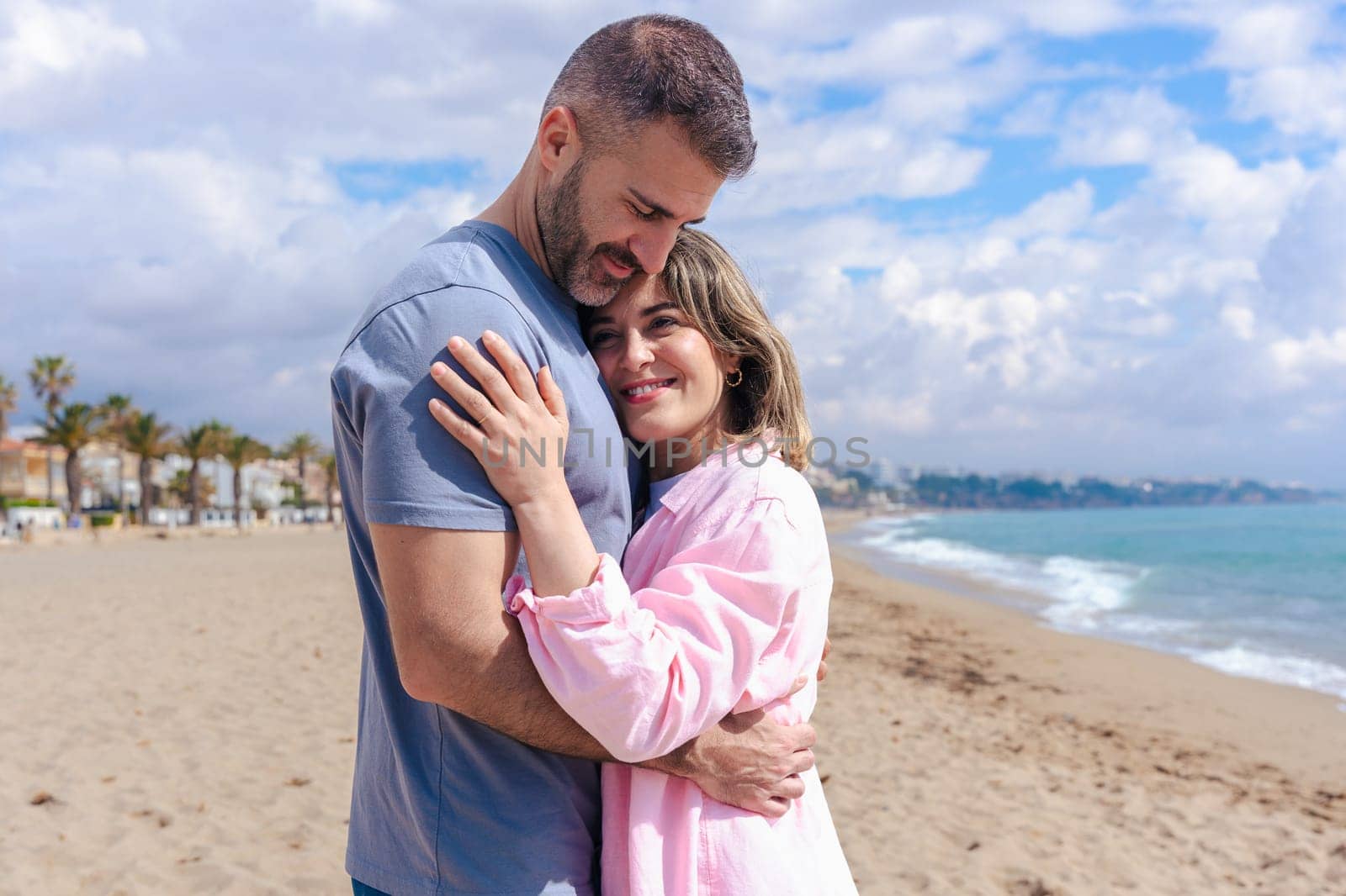 Portrait of a smiling caucasian young couple embracing on a beach during vacation. Relaxed and affectionate moment by the sea, with blue sky, ocean waves, and sandy shore behind them.