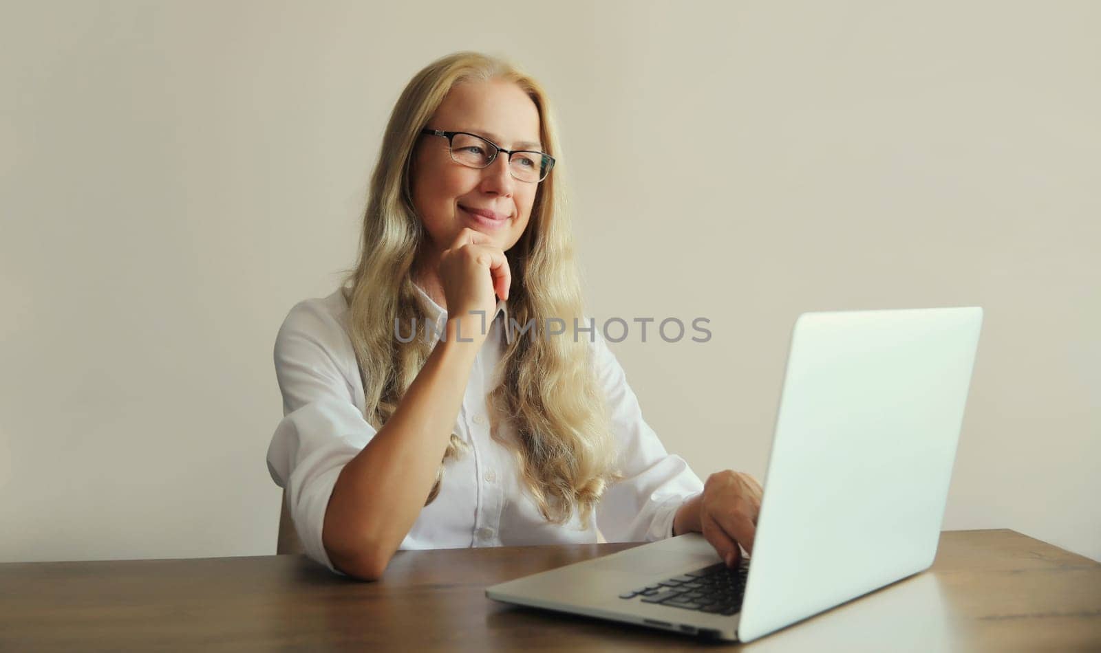 Happy confident mature woman working on laptop wearing a glasses, sitting at desk in office or at home