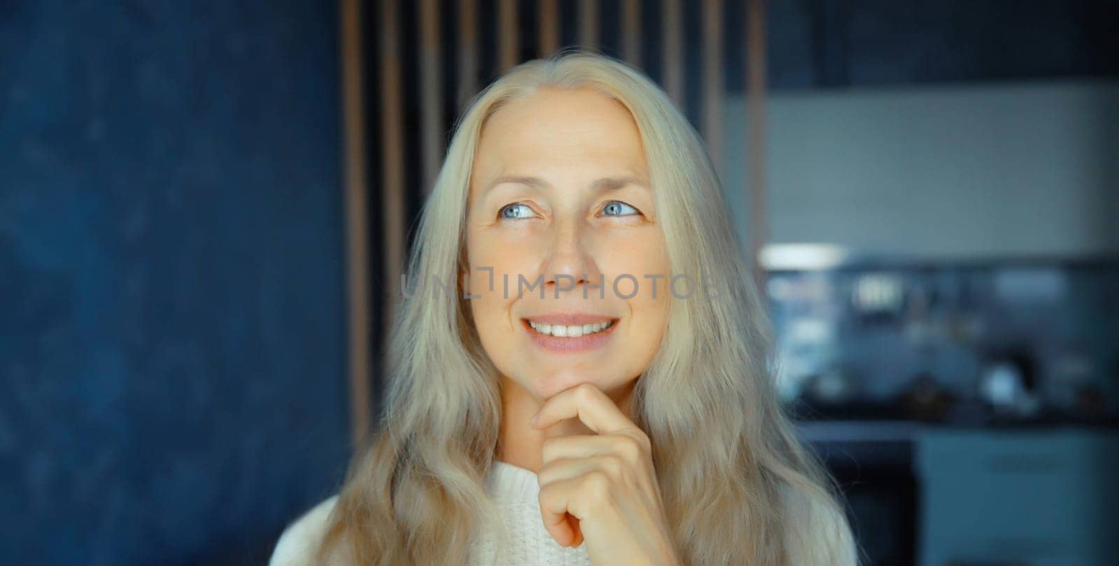 Pensive mature woman thinks looking out the window in early morning in living room with kitchen at home, copy space background