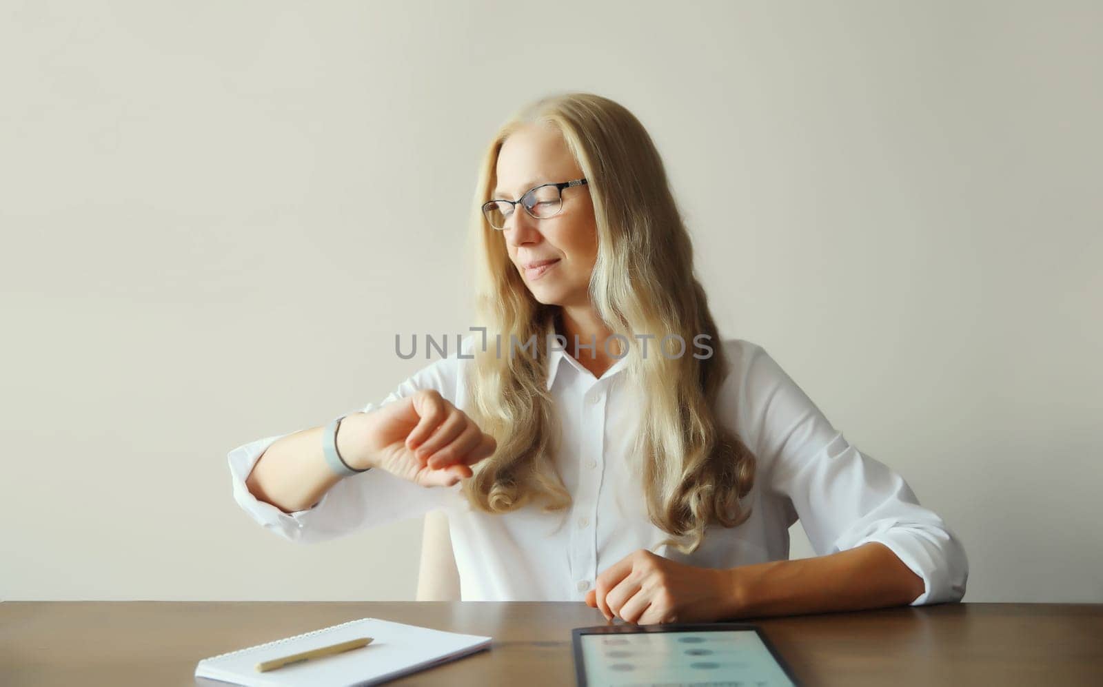 Confident mature woman professional working on digital tablet computer with paper documents looks at watch, checks the time, sitting at desk in office or at home