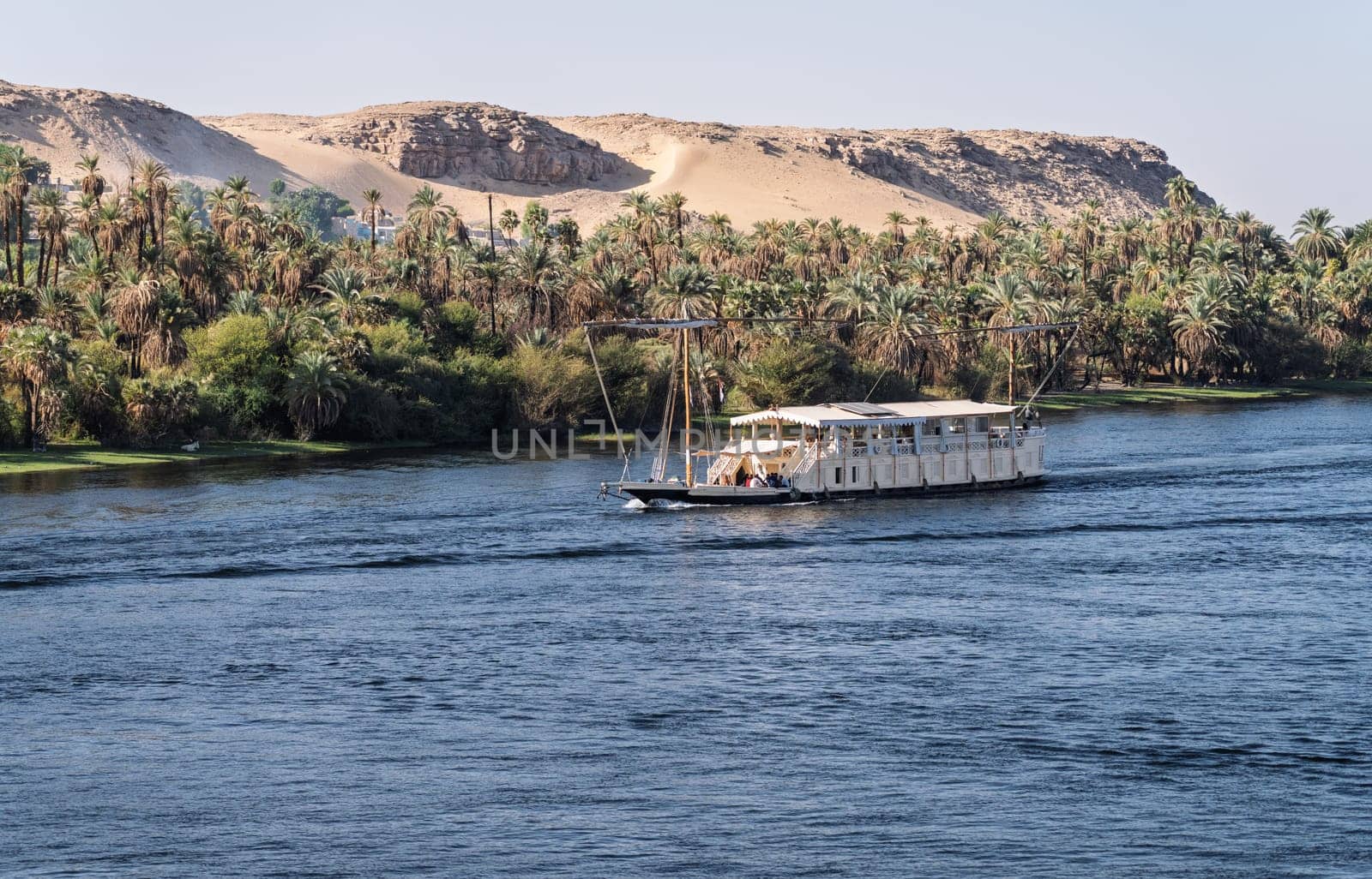 Dahabiya Nile cruise ship sailing alongside a sandy desert with palm trees on the River Nile near Aswan