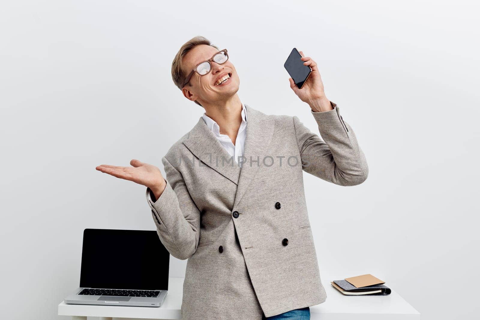 cheerful happy caucasian man talking on smartphone with smile wearing glasses and gray blazer standing near laptop on white background digital nomad remote work concept showing positive feelings
