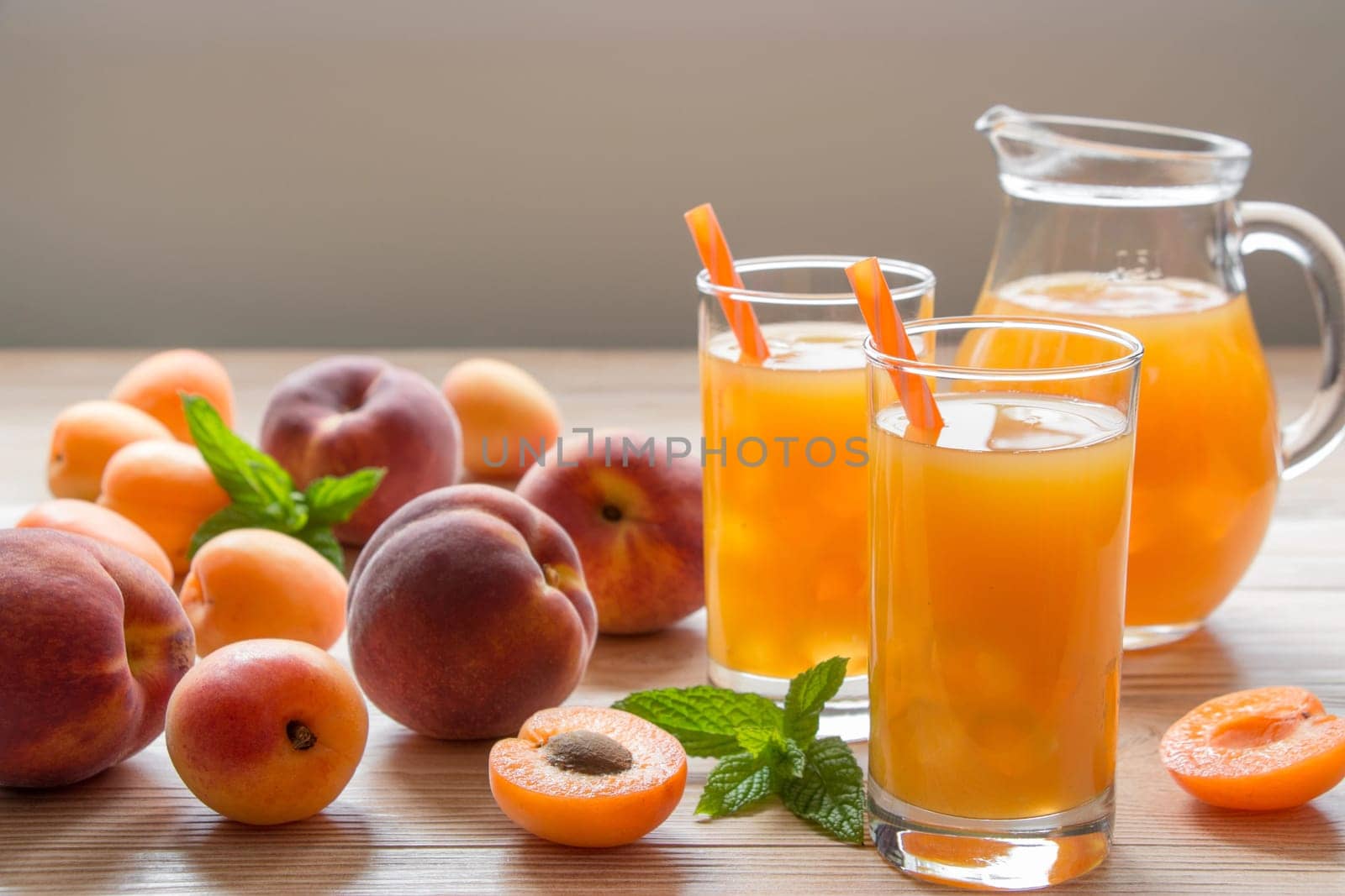 Apricot and peach juice in glasses with ice near the jar of juice and scattered apricots and peaches, mint on a light wooden background. Apricot and peach juice with ice. Horizontal. Daylight. Close.