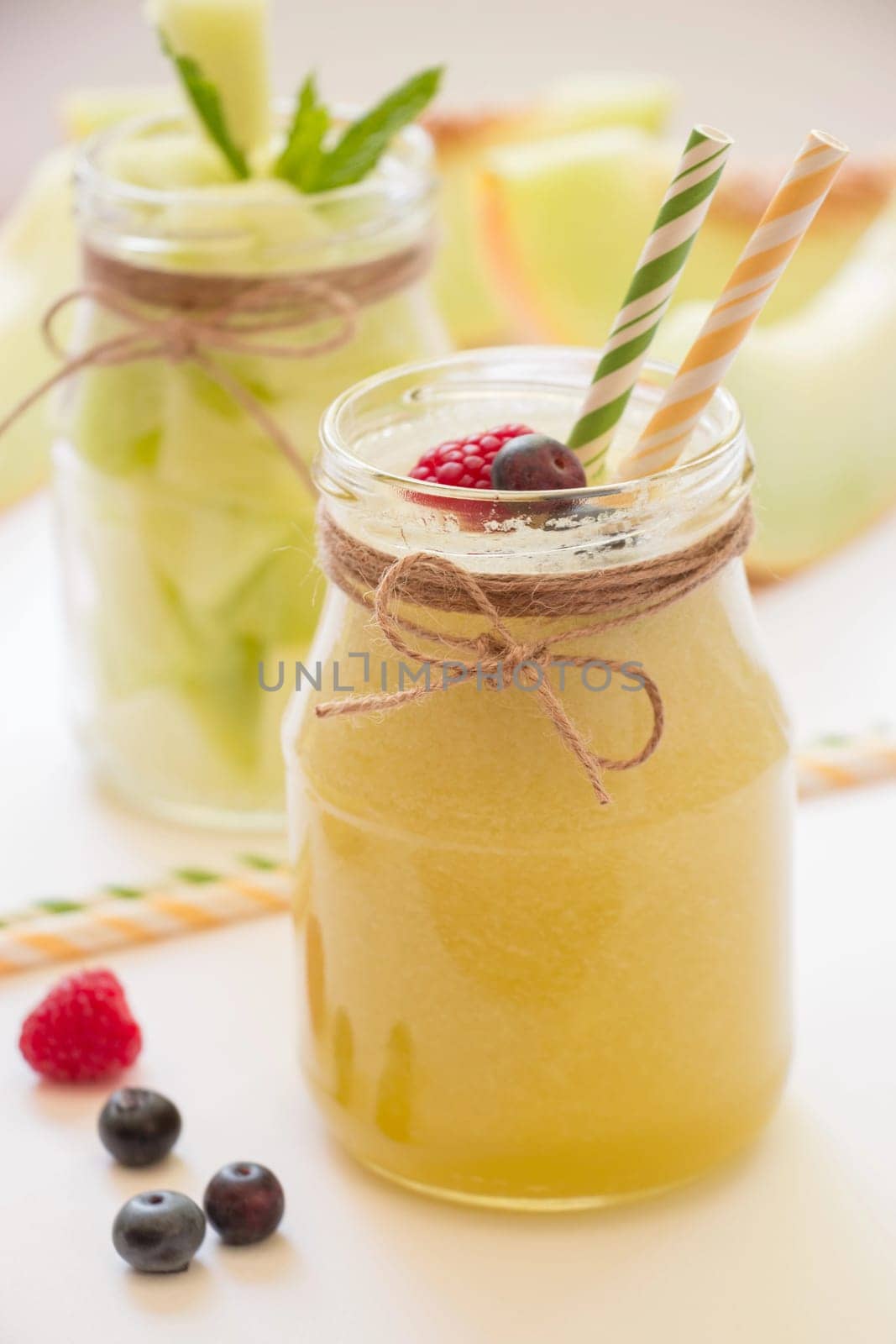 In front a jar with melon smoothie near berries, straws diagonally behind a jar with melon pieces, melon cut slices on a white background. Melon smoothie and salad in 2 jars. Vertical. Close-up.