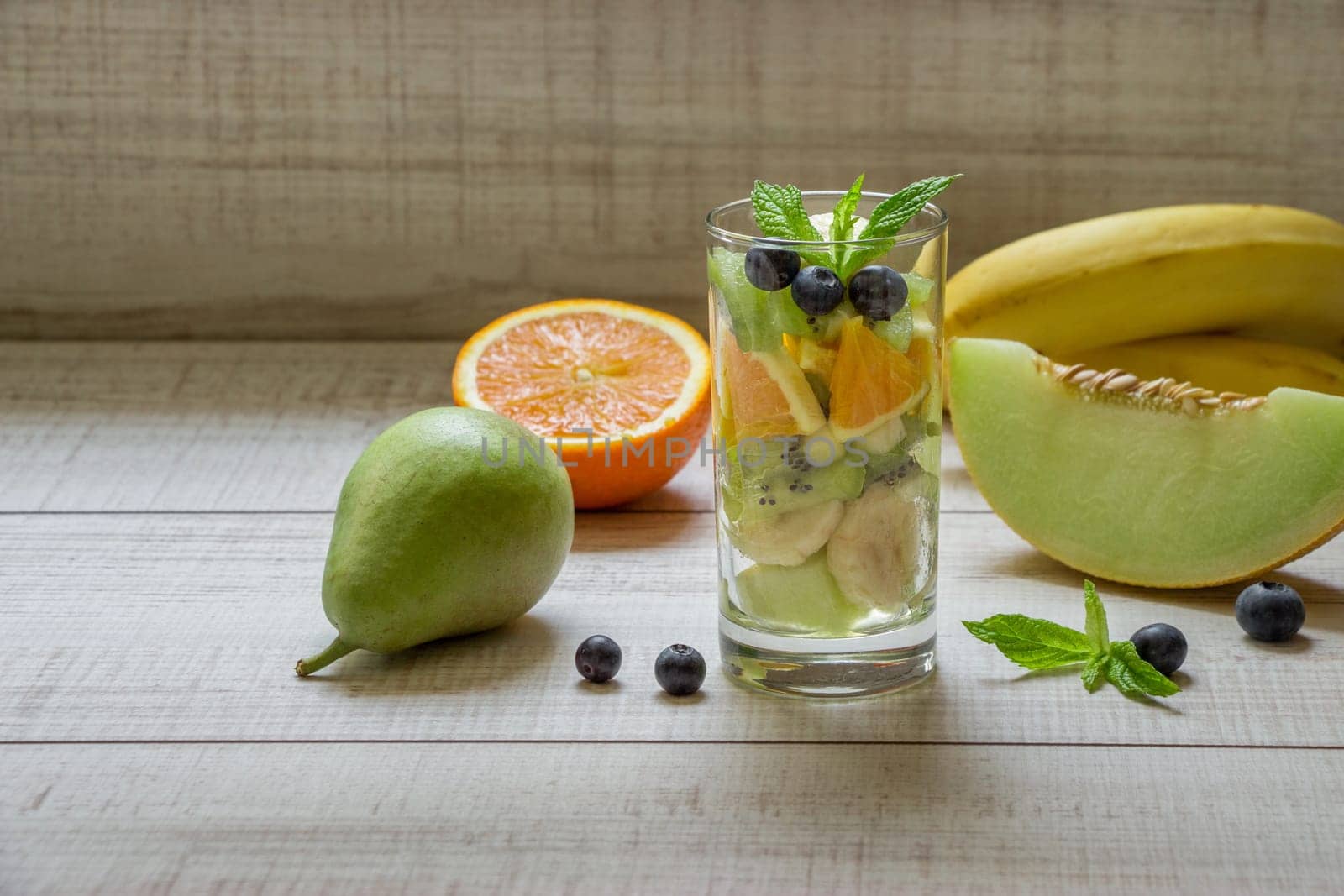 Right glass with sliced fruits melon, banana, orange, kiwi, pear next to ingredients, left empty space for text also in frame blueberries, mint on light background. Glass of fruit salad. Horizontal.