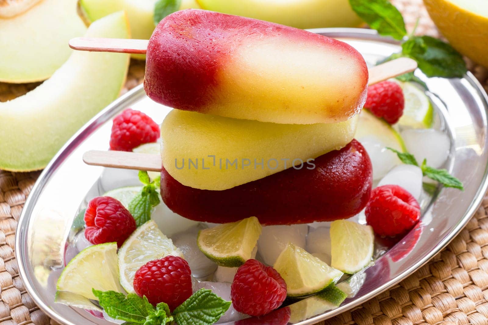 Vibrant melon raspberry ice cream sits atop a mound of ice cubes, surrounded by fresh raspberries, mint leaves, and sliced lime.