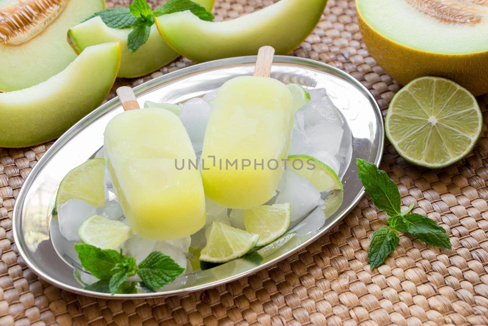 Melon and lime sorbet ice cream on silver plate with ice cubes, lime pieces, mint leaves near melon slices, lime on a straw napkin background. Melon and lime sorbet ice cream popsicles. Horizontal.