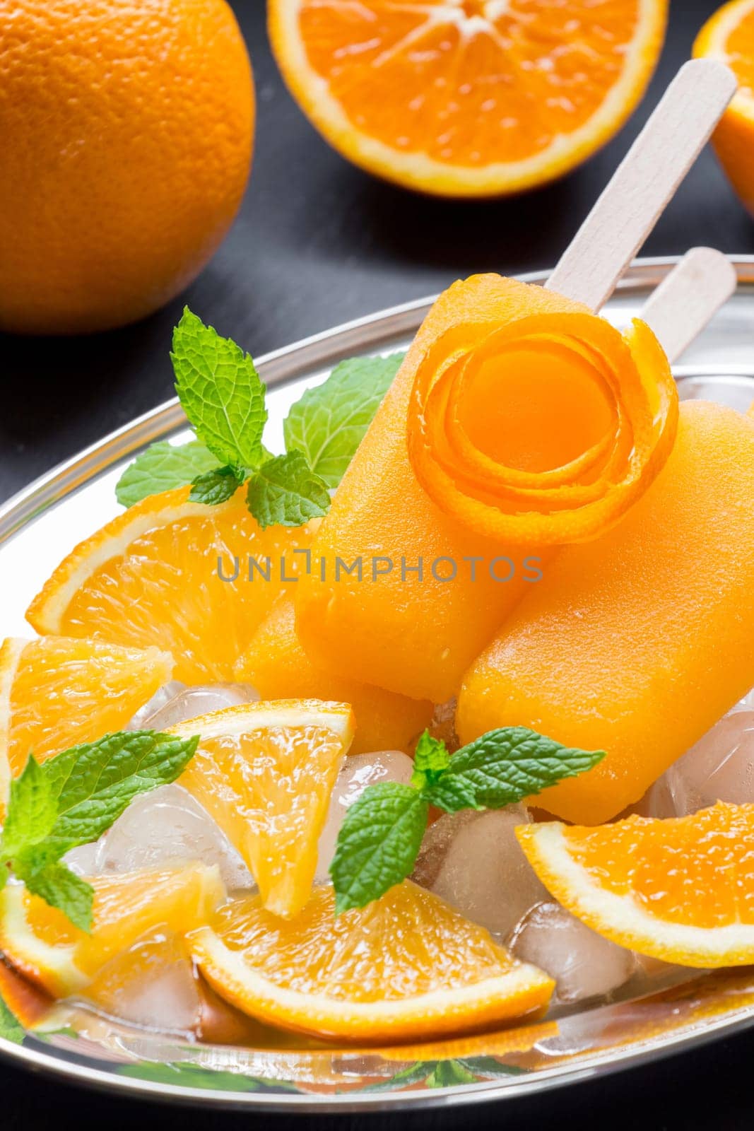 Vibrant orange icecream sits on a silver platter accompanied by fresh orange slices and mint leaves, creating a refreshing dessert perfect for hot summer days.
