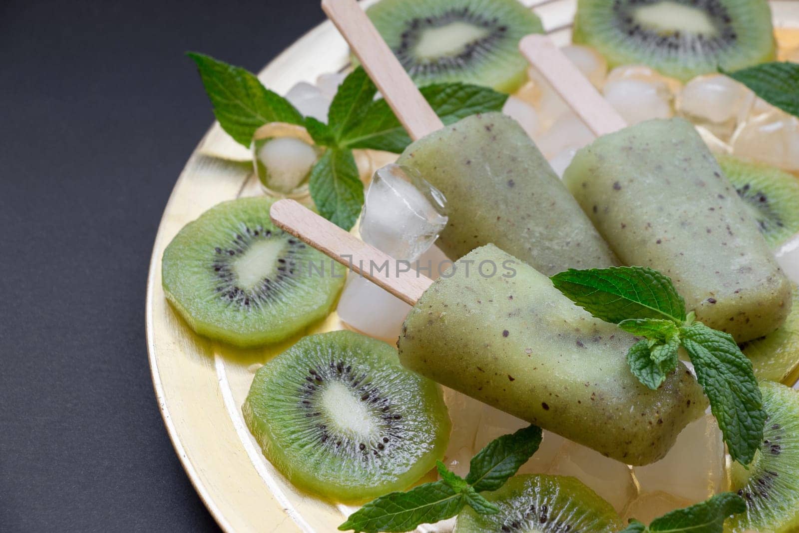 Chilled kiwi icecream popsicles are presented on a golden plate surrounded by ice cubes and fresh kiwi slices, creating a refreshing summer delight at a gathering.