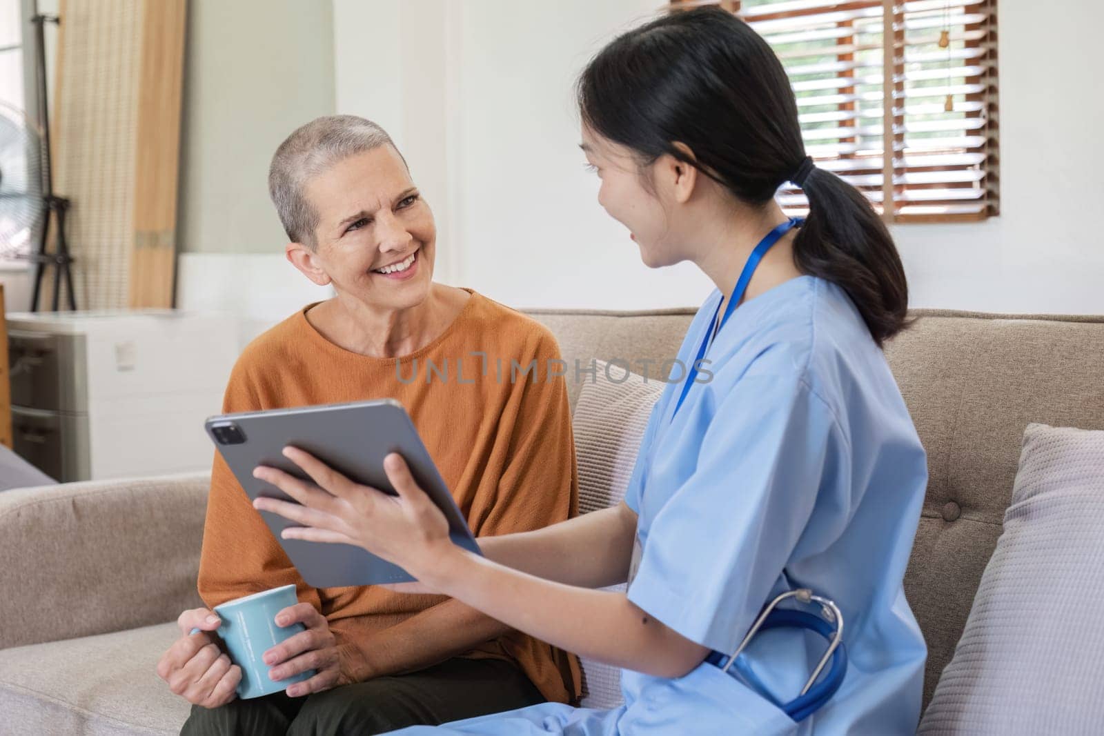 A young nurse offers health care advice to an elderly woman, using a digital tablet in a cozy home environment.
