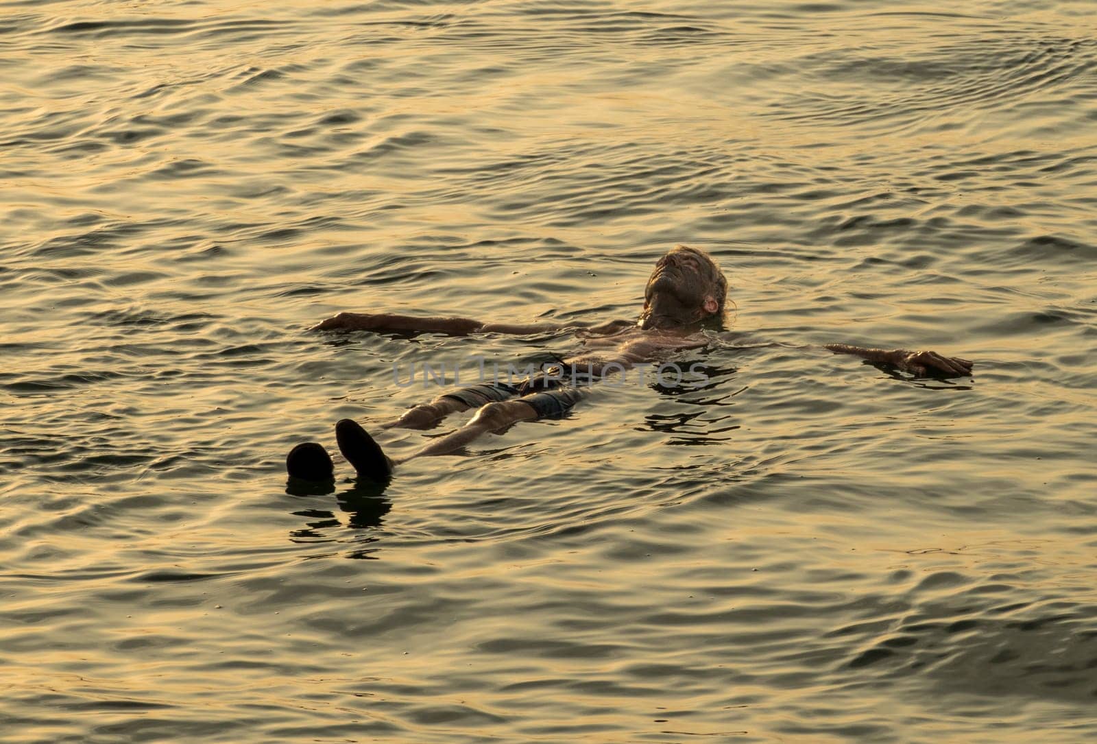 Senior adult man covered with dark mud from the floor of Dead Sea floating in the waters of the salty sea