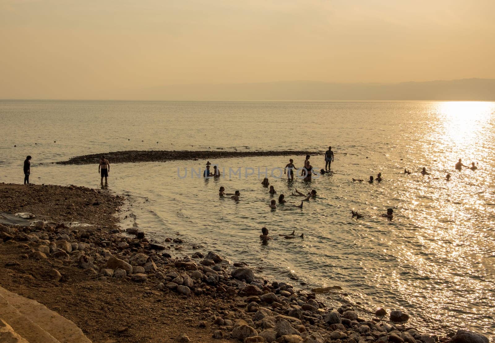 Tourists in the waters of Dead Sea by a beach in modern hotel at sunset