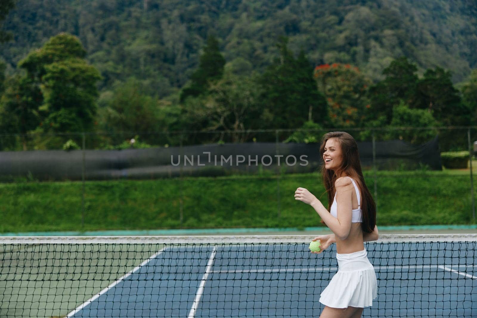 Energetic female athlete in white outfit playing tennis outdoors, exuding joy and health with a beautiful green landscape behind her, ideal for sports and wellness concepts