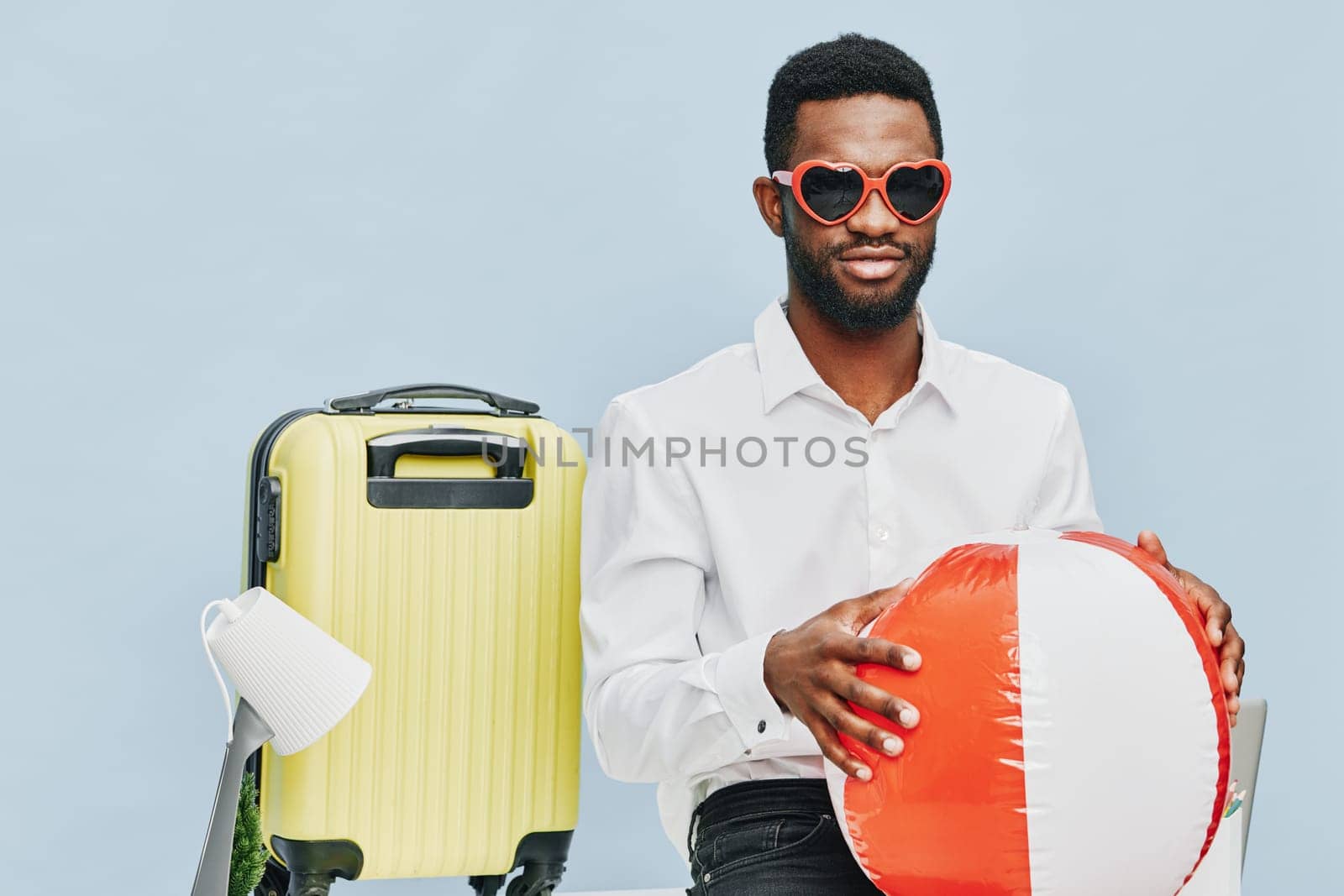 Smiling man with dark skin and glasses, dressed in white shirt, holding inflatable beach ball, standing next to yellow suitcase, studio shot against plain light blue background, summery travel, holiday mood, carefree lifestyle, vacation concept, leisure.