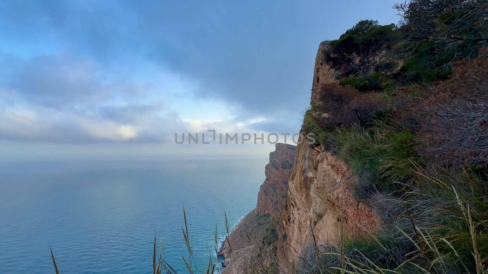 A breathtaking view of a coastal cliff with lush greenery and tranquil ocean waters under a cloudy sky.