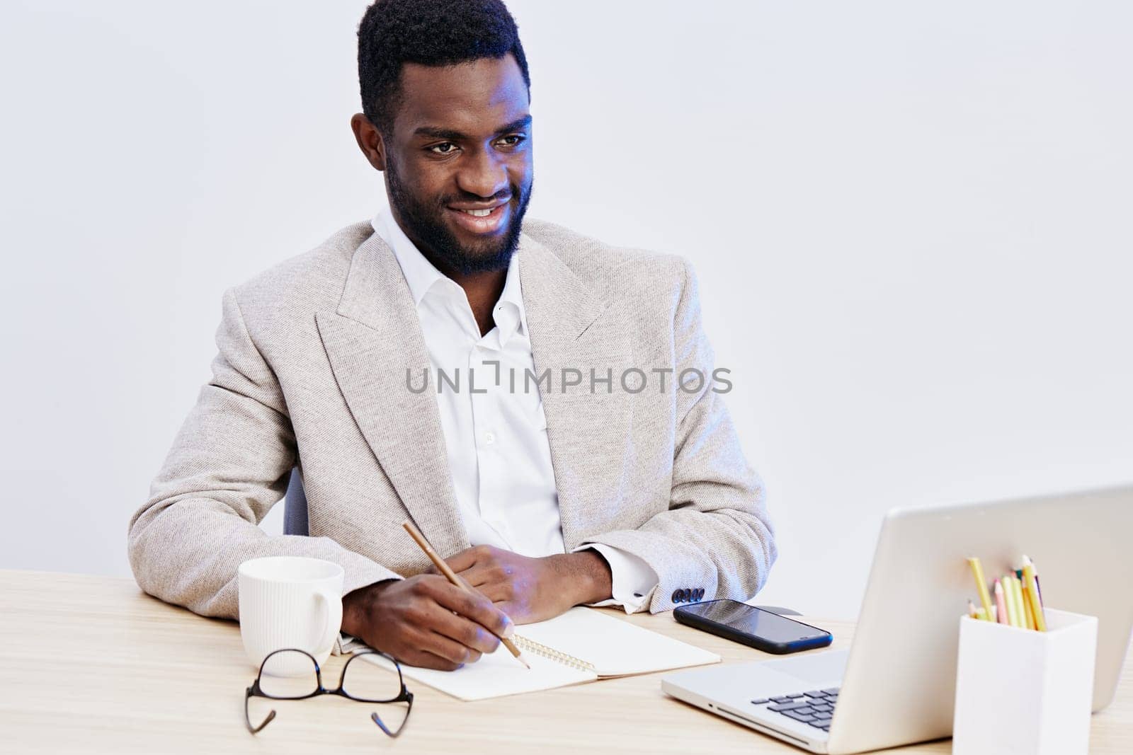 Cheerful young black man dressed in formal attire working confidently in a modern office environment with a laptop, notebook, and writing tools, studio setting, bright and inviting atmosphere, expressing productivity and professionalism, business success.