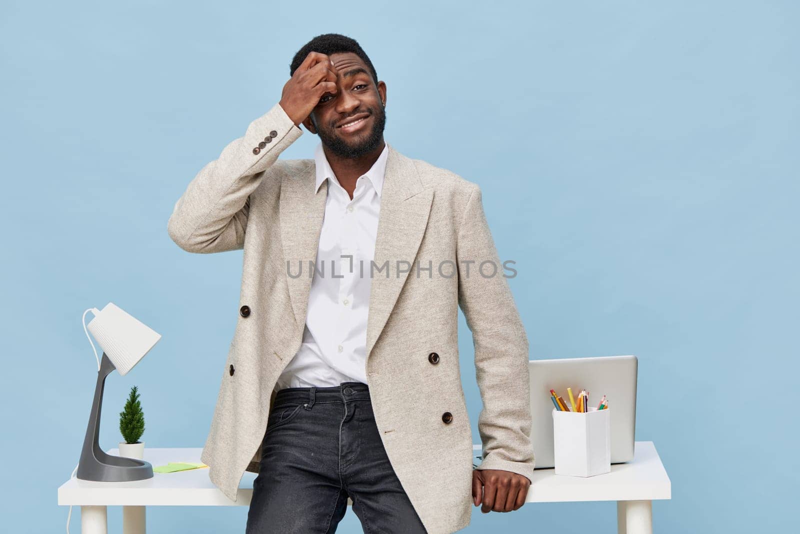 Bright office scene with a cheerful young man with dark skin and short hair, wearing a beige blazer and white shirt, standing at a white desk with laptop and stationery, exhibiting a thoughtful or puzzled expression. Workplace mood concept